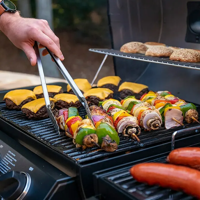 Char-Griller Hybrid-Grill Double Play Gas & Holzkohle - Grillfläche (B x T): 101 x 50 cm, 15,48 kW - Person grillt Burger, Gemüsespieße, Fleischspieße und Hot Dogs auf einem Gasgrill. Brötchen wärmen auf dem oberen Rost.