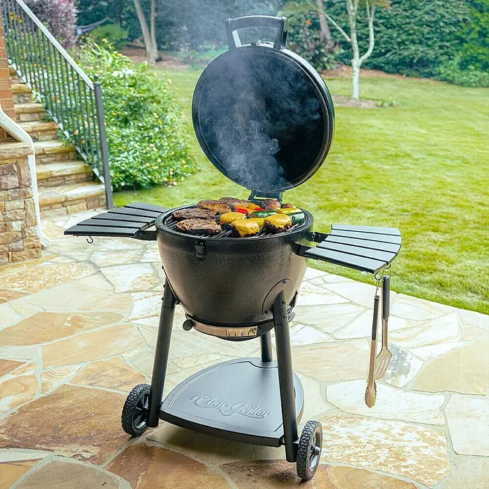 Char-Griller Holzkohlegrill Akorn Kamado - Durchmesser Grillfläche: 50 cm, Schwarz - Schwarzer Grill im Keramik-Stil kocht Burger und Gemüse auf einer Terrasse. Mit Seitenablagen und Rädern.