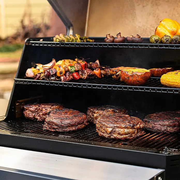 Masterbuilt Holzkohlegrill Gravity 1050 - Grillfläche (B x T): 66 x 49 cm, Schwarz - Ein schwarzer Außengrill mit mehreren Rosten, beladen mit gegrillten Speisen wie Steaks, Maiskolben, Gemüsespießen und gebratenem Kürbis.