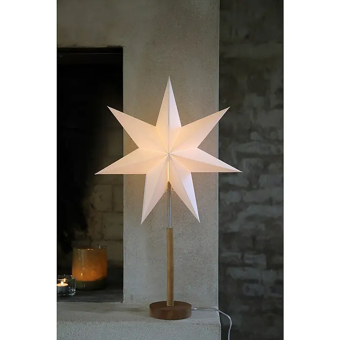 Markslöjd Standstern Speranza - Weiß/ Braun, Netzbetrieben - Beleuchtete, weiße Sternlampe auf Holzfuß steht auf Steinvorsprung neben einem Kamin.