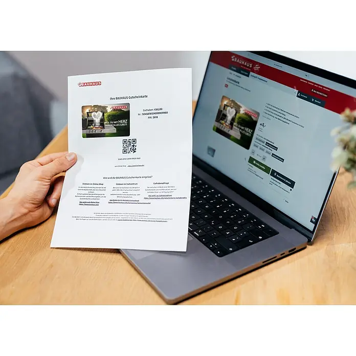 Gutscheinkarte - Zum Selbstausdruck, Individueller Betrag, Mit Herz gegrillt - Bauhaus Gutschein mit €60 Guthaben, gehalten in der Hand, mit Laptop im Hintergrund. Ideal für Heimwerker und Gartenliebhaber.