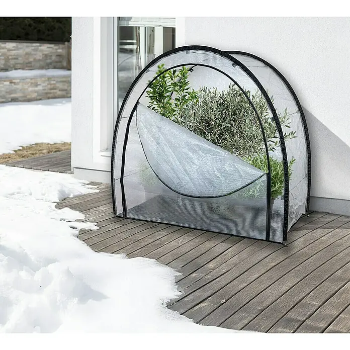Windhager Winterschutzzelt Qinog - 80 x 180 x 170 cm, Gitterfolie - Das Bild zeigt ein kleines, tragbares Gewächshaus mit Pflanzen auf einer Holzterrasse neben einem schneebedeckten Bereich.