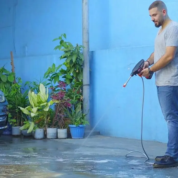 Hidrolimpiadora de alta presión de batería Hydroblast - 9,5 bar, 21 V, Capacidad de bombeo: 210 l/h - Un hombre usa una lavadora a presión portátil para limpiar un área exterior. La herramienta es negra y roja y está removiendo la suciedad.