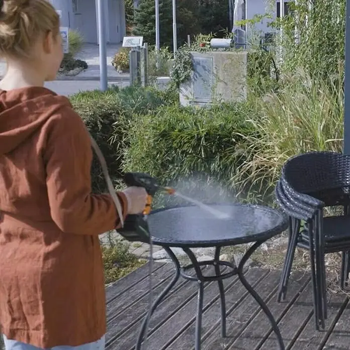 Hidrolimpiadora de alta presión de batería Hydroblast - 9,5 bar, 21 V, Capacidad de bombeo: 210 l/h - Una mujer usa una hidrolavadora para limpiar una mesa de patio negra con tapa de vidrio, rodeada de vegetación y una pila de sillas.