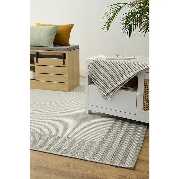 Alfombra exterior In/Out Marco - Gris, 110 x 60 cm, Polipropileno - Una elegante alfombra de rayas grises y blancas se encuentra debajo de un zapatero de madera y un banco con cojines, complementado con una planta.