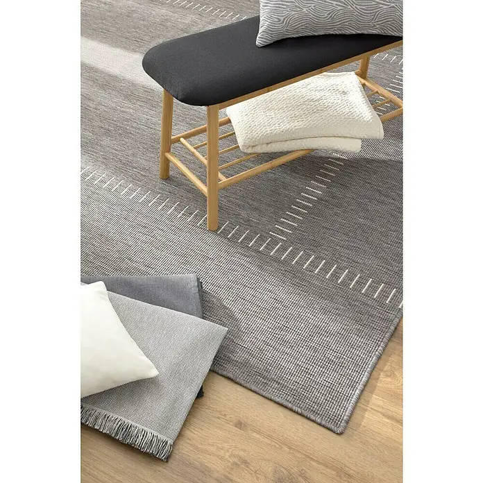 Alfombra exterior reversible In/Out Pespunte - Silver, 230 x 160 cm, Polipropileno - Una alfombra gris con un patrón geométrico, complementada por un banco con cojines y una manta. La alfombra aporta un toque moderno al espacio.