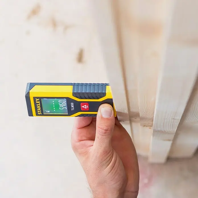 Stanley Medidor de distancia láser TLM50 - Gama de medición: 0,15 m - 15 m - Un medidor láser de distancia Stanley TLM50 se está utilizando para medir una pieza de madera. El dispositivo es amarillo y negro con una pantalla digital.