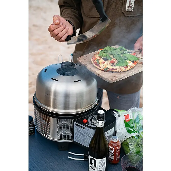 Cobb Gasgrill Premier Gas Deluxe 2.0 - Durchmesser Grillfläche: 32 cm, 1,23 kW, Anzahl Brenner: 1 - Nahaufnahme eines Cobb Grills mit Pizza, Weinflasche und Zutaten auf einem Tisch, während eine Person mit einem Pizzaschneider hantiert.