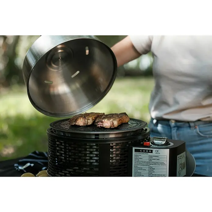 Cobb Gasgrill Pro - Durchmesser Grillfläche: 32 cm, 1,23 kW, Anzahl Brenner: 1 - Ein tragbarer Cobb-Grill mit Steaks darauf, der Deckel wird gerade angehoben, ideal für Picknicks und Camping.