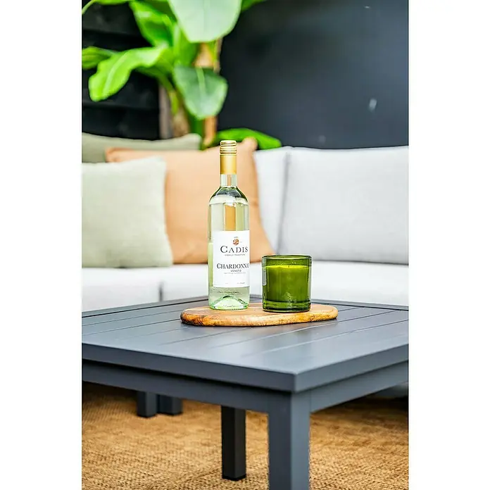 Garden Impressions Loungemöbel-Set Prinos - 4 -tlg., Aluminium, Grau/Schwarz - Eine Flasche Cadis Chardonnay Wein und eine grüne Kerze auf einem Holzbrett, platziert auf einem dunkelgrauen Outdoor-Couchtisch mit Lamellen.