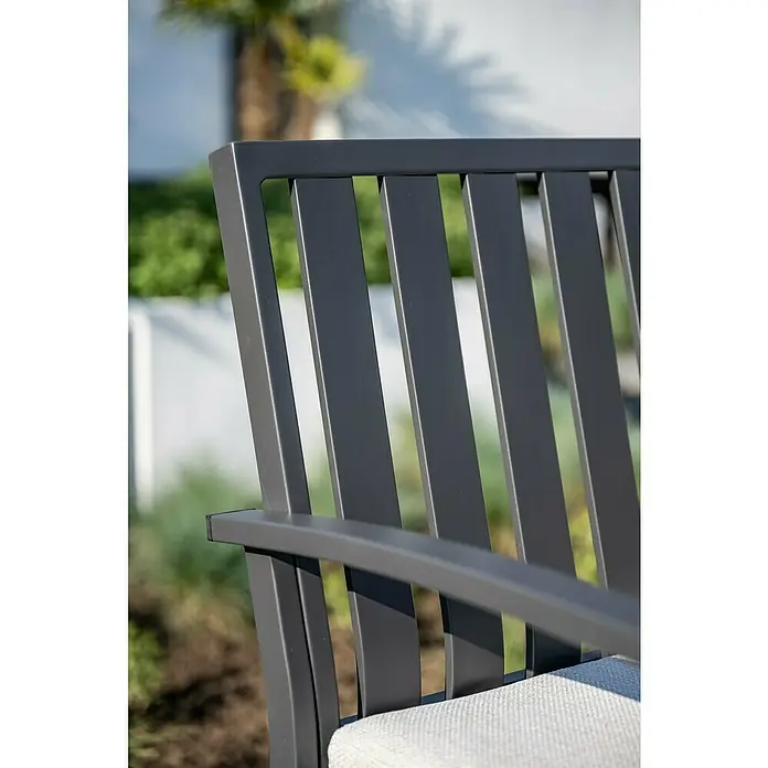 Garden Impressions Gartenbank Pisa - B x T x H: 150 x 52,5 x 89,5 cm, Aluminium, Polyester, Schwarz - Nahaufnahme eines dunkelgrauen Lamellen-Gartenstuhls mit hellgrauem Kissen in einem sonnigen Garten.