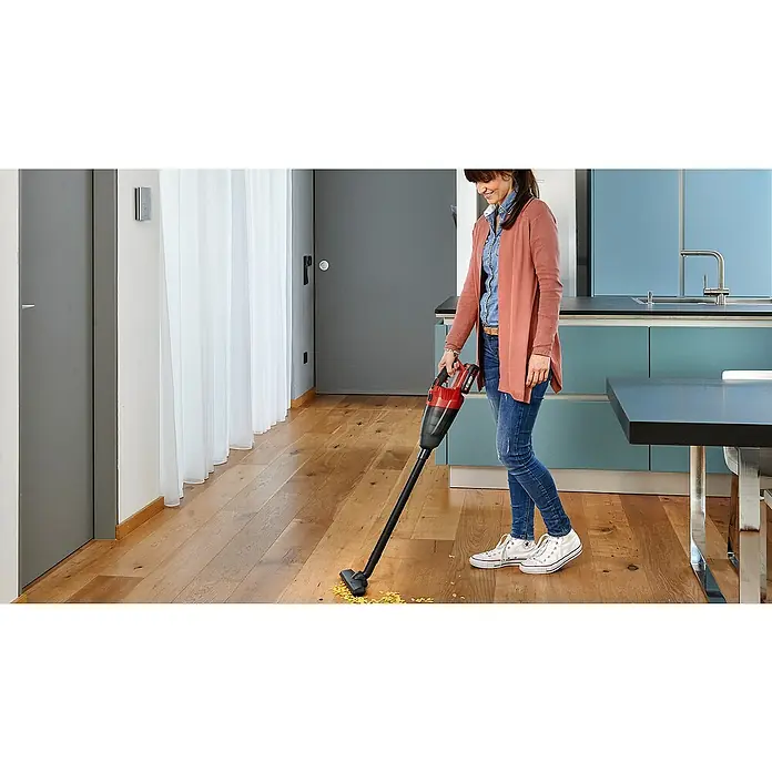 Einhell Power X-Change 18V Akku-Handstaubsauger TE-VC 18 Li-Solo - 18 V, Ohne Akku - Eine Frau staubsaugt Krümel mit einem rot-schwarzen Akku-Staubsauger auf einem Holzboden. Das Gerät ist kompakt und kabellos.