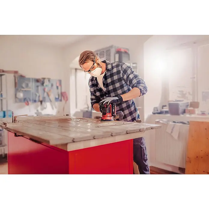 Einhell Power X-Change 18V Akku-Exzenterschleifer TE-RS 18 Li-Solo - 18 V, Ohne Akku, Durchmesser Schleifteller: 125 mm - Ein Mann benutzt einen Einhell Exzenterschleifer zum Schleifen eines Holztisches. Er trägt Schutzbrille, Maske und Handschuhe für die Arbeitssicherheit.