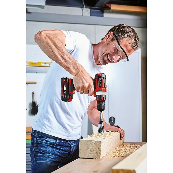 Einhell Power X-Change 18V Akku-Bohrschrauber TE-CD 18/40 Li-Solo - 18 V, Ohne Akku, 40 Nm - Mann bohrt mit einem Einhell Akku-Bohrhammer ein Loch in ein Holzbrett, Holzspäne fliegen. Das Gerät ist rot und schwarz.
