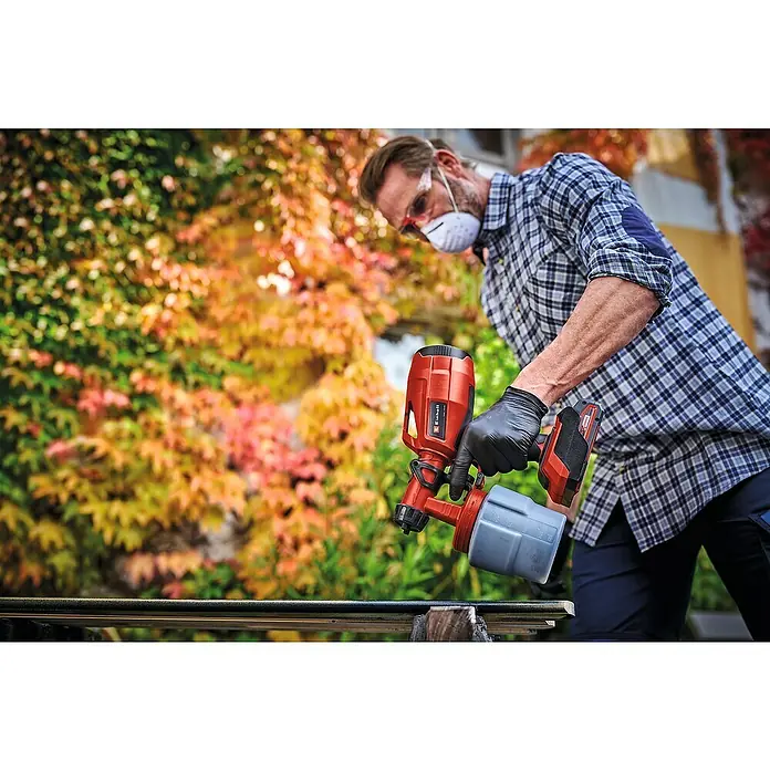 Einhell Power X-Change 18V Akku-Farbsprühsystem TC-SY 18/60 Li-Solo - 18 V, Ohne Akku - Ein Mann verwendet eine Einhell Akku-Farbspritzpistole, um ein Metallstück zu lackieren, vor einem Hintergrund aus bunten Herbstblättern.