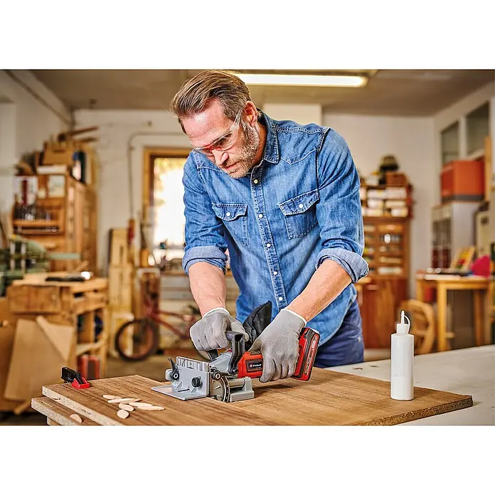 Einhell Power X-Change 18V Akku-Flachdübelfräse TE-BJ 18 Li - Solo - 18 V, Ohne Akku, Leerlaufdrehzahl: 6.500 U/min - Mann arbeitet mit einer Einhell Akku-Nutfräse an einem Holzbrett, um es zu bearbeiten. Er trägt dabei Schutzbrille und Handschuhe.