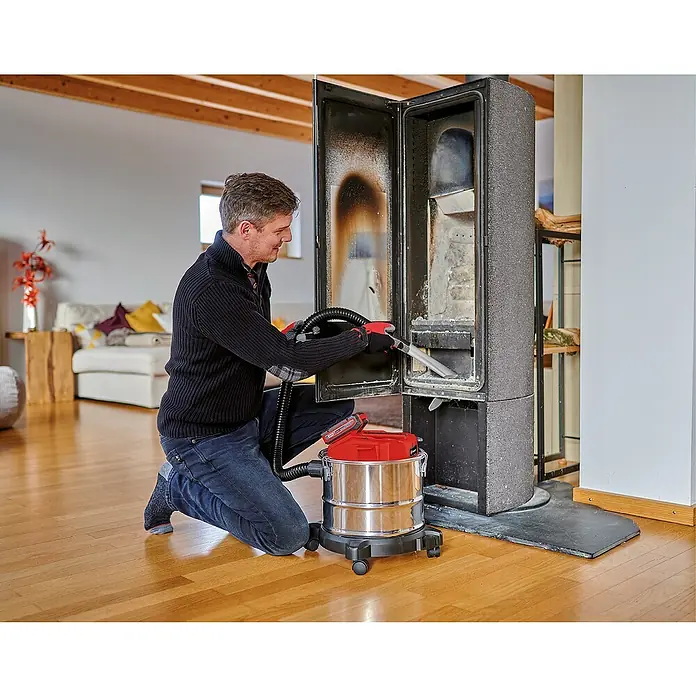 Einhell Power X-Change 18V Akku-Aschesauger TE-AV 18/15 LI-C Solo - 18 V, Li-Ionen, Ohne Akku - Ein Mann reinigt einen Kaminofen mit einem Aschesauger. Der Sauger steht auf Rollen und hat einen roten Deckel. Der Mann trägt Handschuhe.