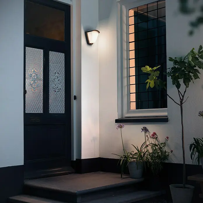 Philips Aplique exterior KisKadee - 42 W, 197 x 140 x 212 mm, Antracita/Blanco, IP44 - Una elegante lámpara de pared exterior ilumina con estilo la entrada con puerta y ventana, rodeada de plantas.
