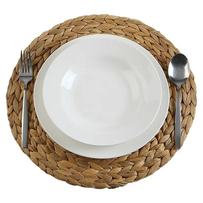 Versa Salvamantel Redondo - Diámetro: 37 cm, Marrón - Elegante decoración de mesa con un plato blanco sobre un individual redondo de fibra natural, acompañado de un tenedor y una cuchara.