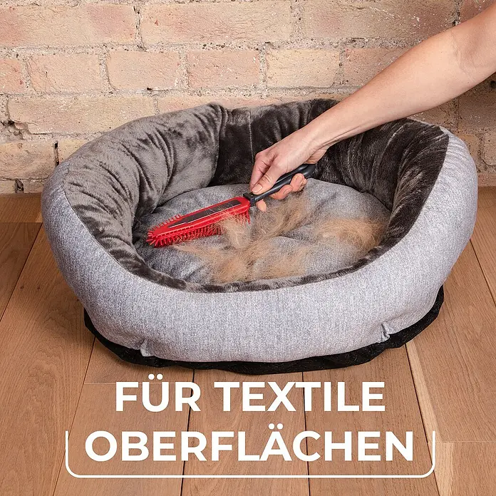 Vileda pet pro Tierhaar-Entferner Electrostatic Brush - Rot/Schwarz, Material Borsten: Gummi, Kunststoff - Eine Hand bürstet Tierhaare mit einer Fusselbürste von einem Hundebett. Die Bürste entfernt effektiv Haare von Textiloberflächen.