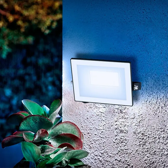 Philips Proyector LED Lois Flood - 20 W, Negro, L x An x Al: 2 x 13 x 11 cm, Blanco frío - Un elegante foco LED de exterior ilumina una pared texturizada, proporcionando seguridad y ambiente en el jardín.