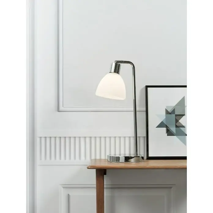 Nordlux Ray Lámpara de sobremesa - 40 W, L x An x Al: 12 x 12 x 46 cm, Blanco, E14 - Lámpara de escritorio de cromo pulido con pantalla blanca, sobre una mesa de madera con un cuadro enmarcado detrás.