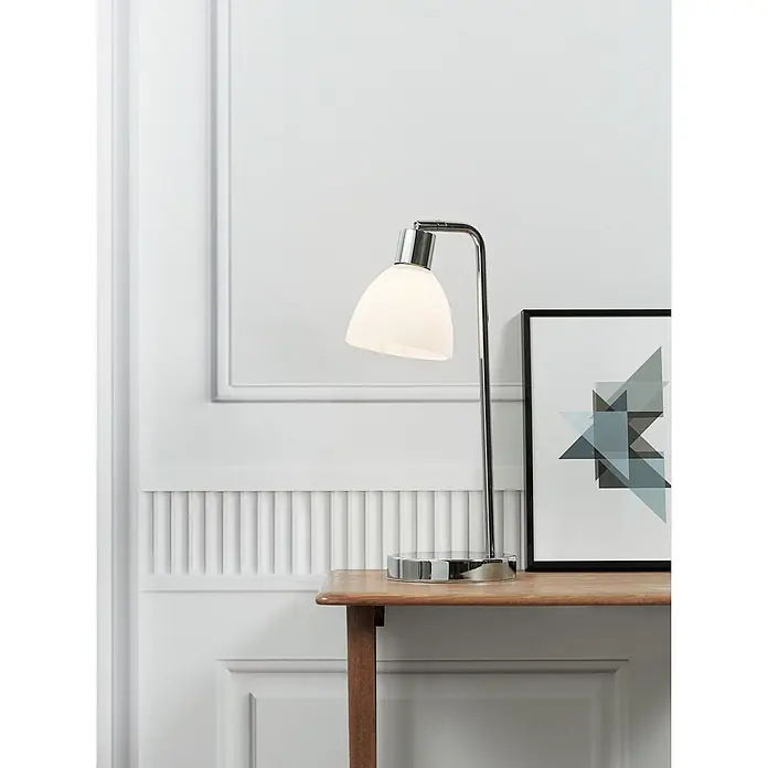 Nordlux Ray Lámpara de sobremesa - 40 W, L x An x Al: 12 x 12 x 46 cm, Blanco, E14 - Lámpara de escritorio de cromo pulido con pantalla blanca, sobre una mesa de madera con un cuadro enmarcado detrás.
