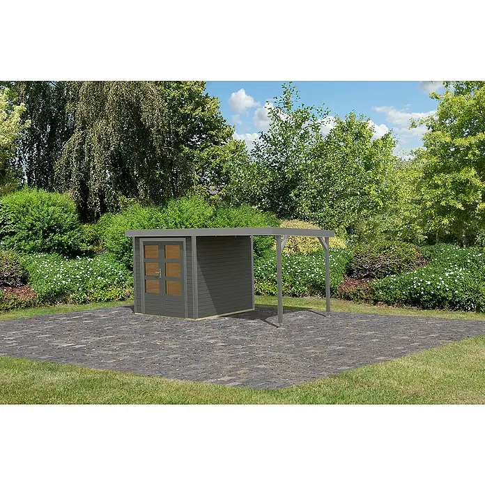 Karibu Gartenhaus Jupiter 2 B - Außenmaß inkl. Dachüberstand (B x T): 453 x 235 cm, Holz, Terragrau/Wassergrau - Abbildung eines Gartenhauses mit angebautem Unterstand auf einer gepflasterten Fläche, umgeben von Grünpflanzen und Bäumen.