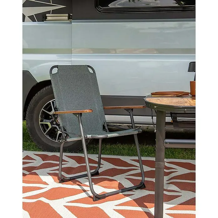 Bo-Camp Campingstuhl Bushwick M - Aluminium, Bambus, Grün, B x T x H: 52 x 60 x 90 cm - Ein grauer Faltstuhl mit Holzarmlehnen steht auf einem gemusterten Teppich, neben einem Tisch und vor einem Wohnmobil.
