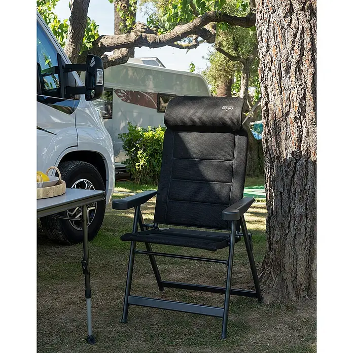 Crespo Campingstuhl Air-Deluxe M - B x T x H: 69 x 74 x 119 cm, Schwarz, Aluminium, Meshgewebe - Ein schwarzer Campingstuhl mit Kopfstütze steht neben einem Tisch und einem Baum im Freien. Ideal für entspannte Momente beim Camping.