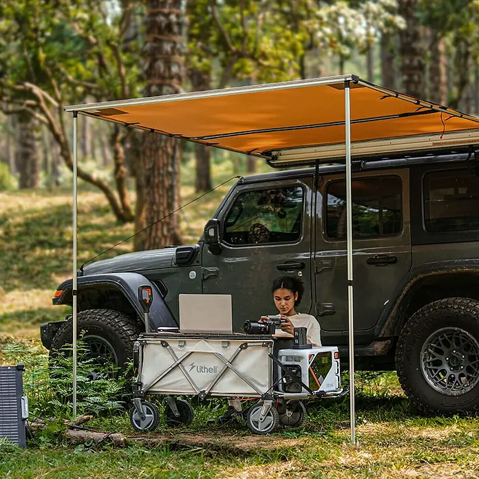 Litheli Bollerwagen 450 W W1 Pro EHW030005 -  - Eine Frau arbeitet an einem Laptop im Freien neben einem Litheli-Campingwagen und einem dunklen Geländewagen.
