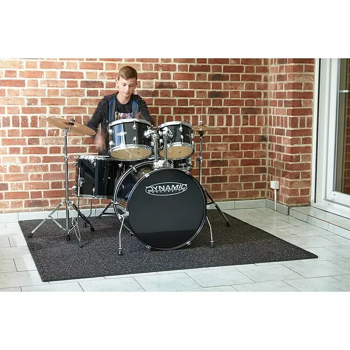 Mottez Estera antideslizante ensamblable B516VPRO - Negro, L x An: 62 x 62 cm - Un baterista adolescente toca una batería Dynamic Percussion sobre una alfombra negra frente a una pared de ladrillos.