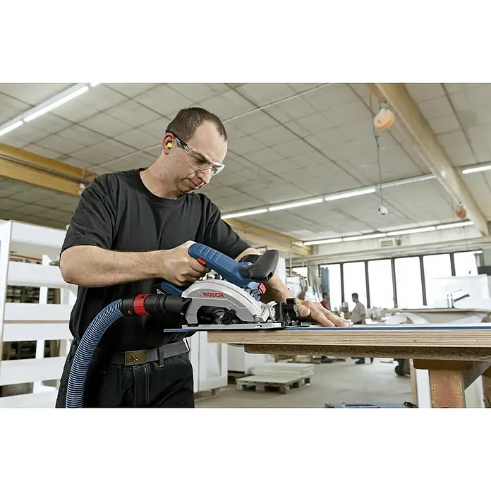 Bosch Professional AMPShare 18V Akku-Handkreissäge GKS 18V-57 G L-Boxx - 18 V, Ohne Akku, Leerlaufdrehzahl: 3.400 U/min, Sägeblatt: 165 mm - Ein Handwerker, der mit einer Bosch Tauchsäge ein Holzbrett bearbeitet. Die Säge ist mit einem blauen Schlauch verbunden und gleitet auf einer Führungsschiene.