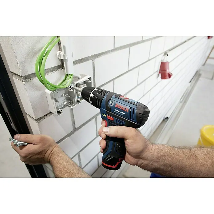 Bosch Professional Akku-Bohrschrauber GSR 12V-15 - 12 V, 2 Akkus, 2 Ah, 30 Nm, Ladegerät + Tasche - Ein Mann befestigt eine graue Anschlussdose mit einem blauen Bosch GSB 10,8-2-LI Akku-Bohrschrauber an einer Ziegelwand.