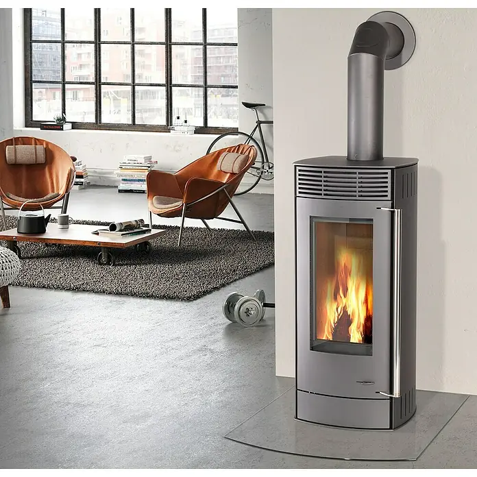 Kaminofen Alpha - 6 kW, Raumheizvermögen: 172 m³, Material Verkleidung: Stahl, Farbe Verkleidung: Gussgrau - Modernes Wohnzimmer mit Kaminofen, der Wärme und Gemütlichkeit ausstrahlt. Der Ofen steht auf einer Glasplatte vor einer weißen Wand.