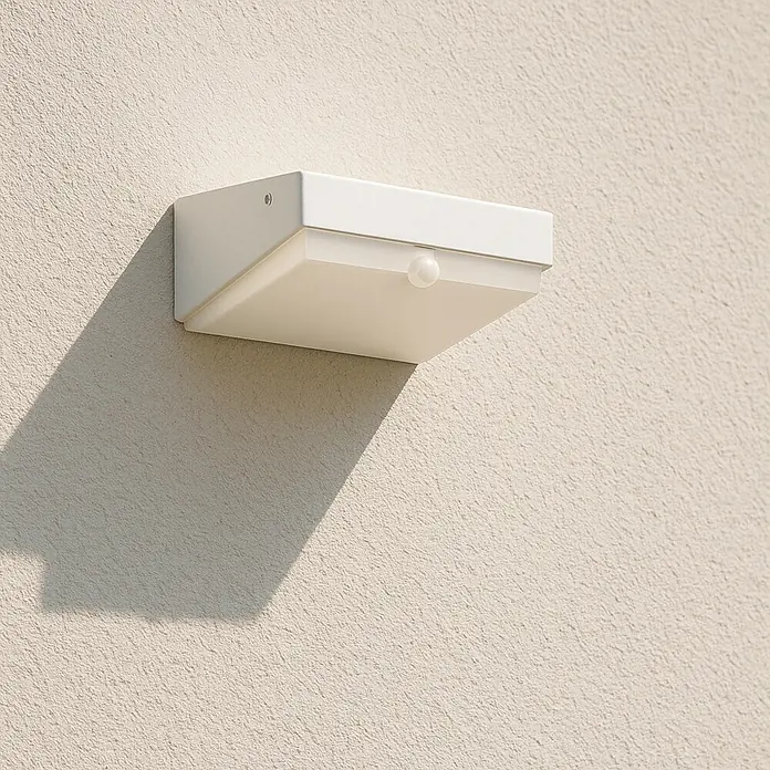 Arte confort Aplique solar para exterior con sensor Alcor - 5,6 W, L x An x Al: 18 x 9 x 4,5 cm, Plateado - Una luz exterior blanca y rectangular con sensor de movimiento está montada en una pared beige, proyectando una sombra marcada.