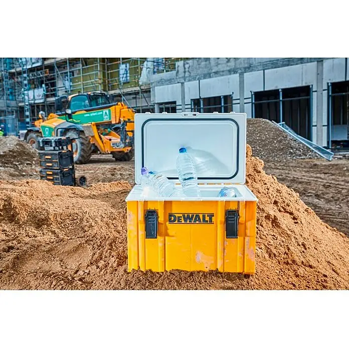 DeWalt Nevera portátil DWST1-81333 - L x An x Al: 36,5 x 55,5 x 40,8 cm - La nevera amarilla DeWalt con botellas de agua se encuentra en una colina de arena, con una obra y equipos de construcción al fondo.