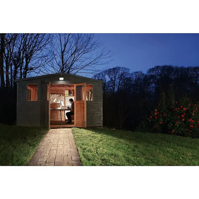 Ledvance Essential LED-Strahler - 30 W, 2.850 lm, 4.000 K, L x B x H: 186 x 128 x 28 mm, IP65 - Außenaufnahme eines Arbeitsplatzes in einem Gartenhaus bei Nacht, beleuchtet, mit Person am Schreibtisch.