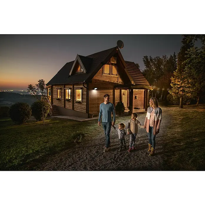 Osram LED-Strahler Floodlight Essential Sensor - 10 W, L x B x H: 13,2 x 8,3 x 3,3 cm, Schwarz, Tageslichtweiß, Mit Sensor - Eine Familie spaziert vor ihrem beleuchteten Holzhaus. Die Außenbeleuchtung betont das gemütliche Ambiente und sorgt für Sicherheit.