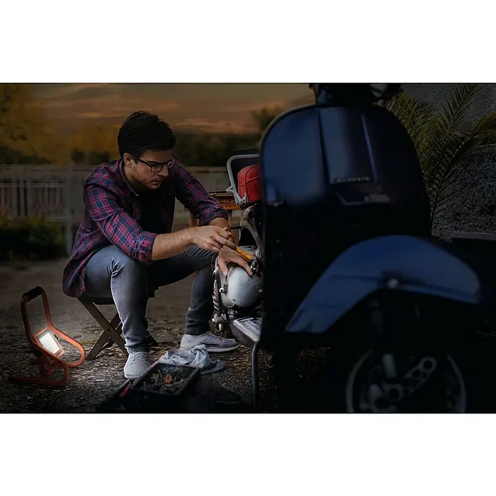 Ledvance LED-Baustrahler R-Stand - 20 W, 1.800 lm, 4.000 K, 266 x 200 x 276 mm, IP44 - Orangefarbene, tragbare LED-Arbeitsleuchte beleuchtet einen Mann, der abends einen Roller repariert.
