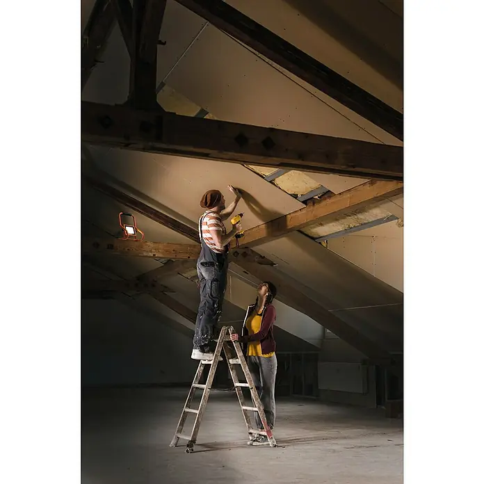 Ledvance LED-Baustrahler R-Stand - 20 W, 1.800 lm, 4.000 K, 266 x 200 x 276 mm, IP44 - Eine Person auf einer Holzleiter montiert mit einem gelben Bohrer eine Deckenplatte im Dachboden, von einer anderen gestützt.