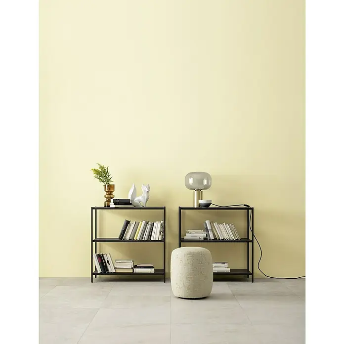 SCHÖNER WOHNEN-Farbe Wandfarbe Designfarbe - Belebendes Champagnercreme, Matt, 2,5 l, Konservierungsmittelfrei - Bücherregale aus schwarzem Metall mit dekorativen Elementen und einem grauen Hocker vor einer hellgelben Wand.