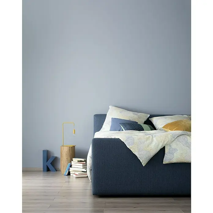 SCHÖNER WOHNEN-Farbe Wandfarbe Designfarbe - Entspanntes Nordischblau, Matt, 2,5 l, Konservierungsmittelfrei - Gemütliches Schlafzimmer-Setup mit einem stilvollen, marineblauen Polsterbett, akzentuiert durch dekorative Kissen und einer Nachttischlampe.