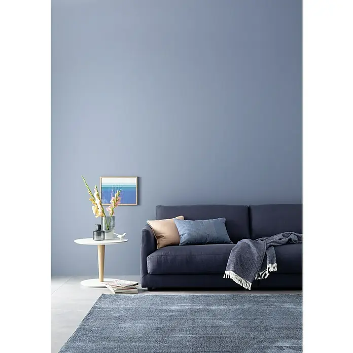 SCHÖNER WOHNEN-Farbe Wandfarbe Designfarbe - Inspirierendes Horizontblau, Matt, 2,5 l, Konservierungsmittelfrei - Stilvolles Wohnzimmer mit dunkelblauem Sofa, passendem Teppich und einem Beistelltisch mit Blumenvase und Dekorationen.