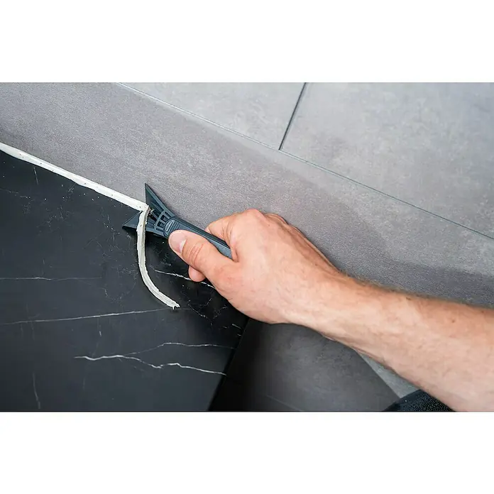 Wolfcraft Rascador de juntas - 4 pzs. - Una mano usa un rascador de juntas para quitar silicona vieja entre baldosas, facilitando la renovación del baño.