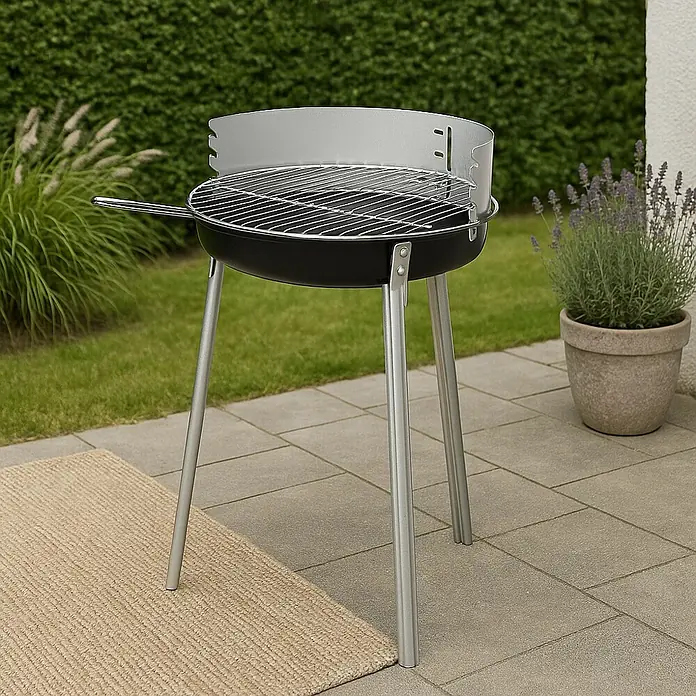 Grillstar Barbacoa de carbón Denver - Diámetro de la superficie de la parrilla: 34 cm, Negro/Plateado - Una barbacoa portátil de carbón con tres patas en un patio, rodeada de vegetación. Perfecta para parrilladas al aire libre.