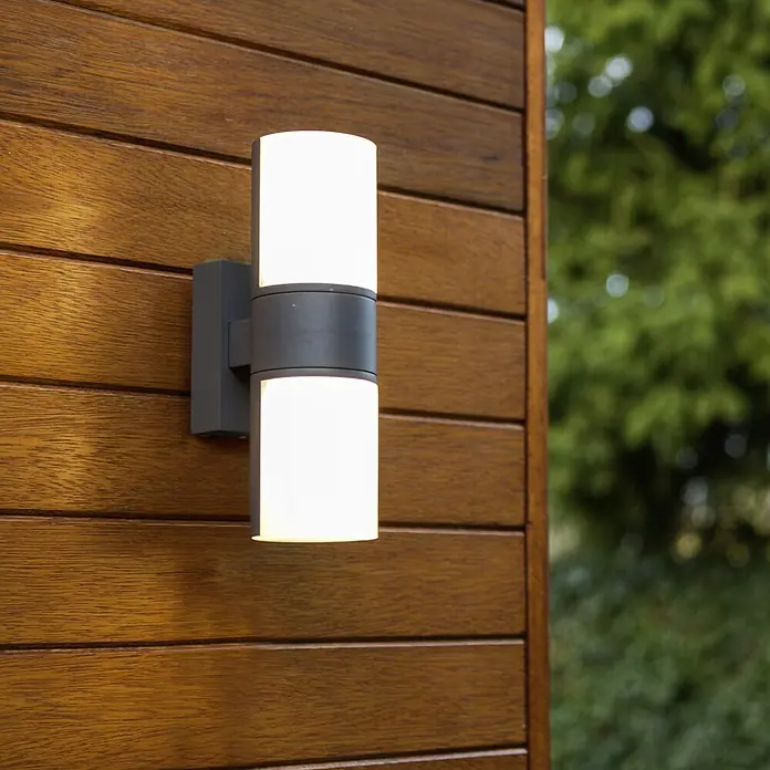 Lutec LED-Außenwandleuchte Cyra - 15 W, 7,8 x 12 x 24,5 cm, Anthrazit/Weiß, IP54 - Nahaufnahme einer modernen Außenwandleuchte mit einem zylindrischen Design auf einer Holzwand.