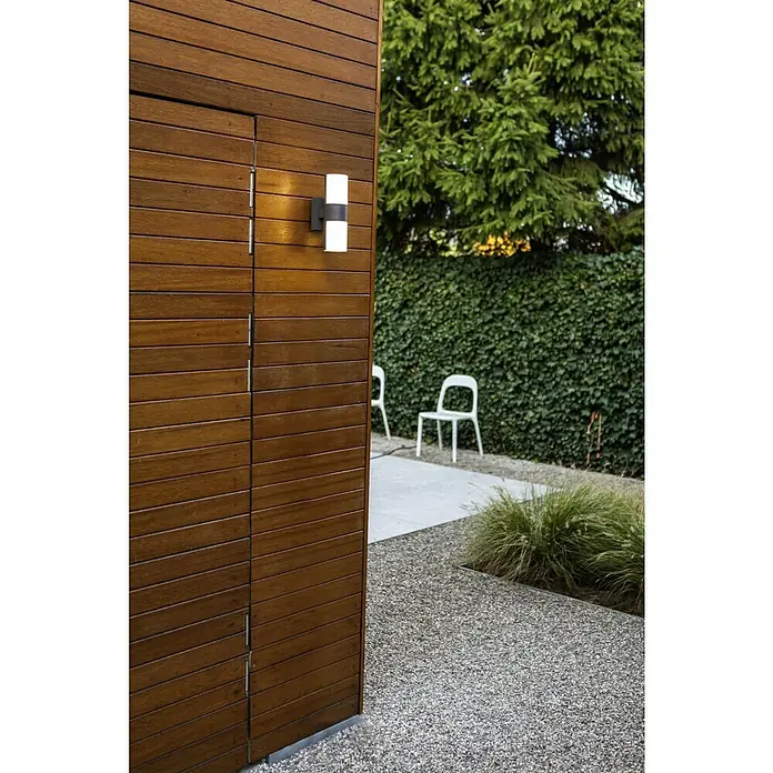 Lutec LED-Außenwandleuchte Cyra - 15 W, 7,8 x 12 x 24,5 cm, Anthrazit/Weiß, IP54 - Eine moderne Außenwandleuchte mit zylindrischem Design beleuchtet eine Holzwand neben einem Garten mit Stühlen und Grünflächen.