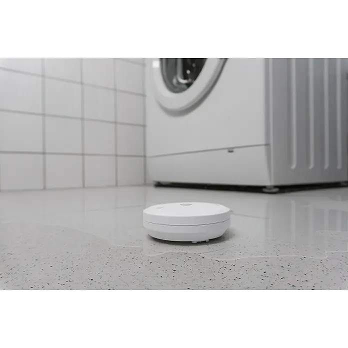 Homematic IP Wassersensor HmIP-SWD - Weiß, Ø x H: 8 x 3 cm, Batteriebetrieben - Das Bild zeigt einen runden, weißen Wassersensor auf einem gefliesten Boden in der Nähe einer Waschmaschine. Der Sensor detektiert Wasseraustritte.