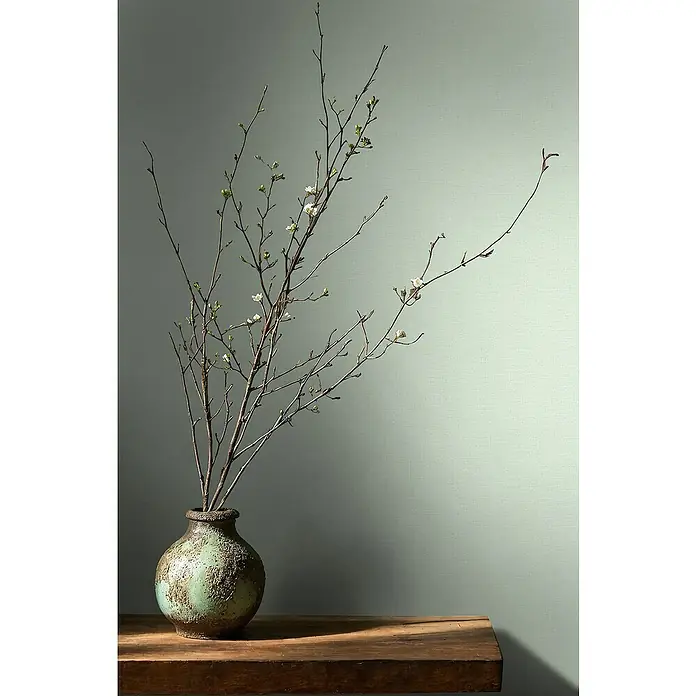 Glööckler Pompöös Provence Vliestapete - Pastellgrün, Uni, 10,05 x 0,53 m - Nahaufnahme einer rustikalen grünen Vase mit zarten Zweigen und Knospen auf einem dunklen Holztisch, vor einem hellgrünen Hintergrund.